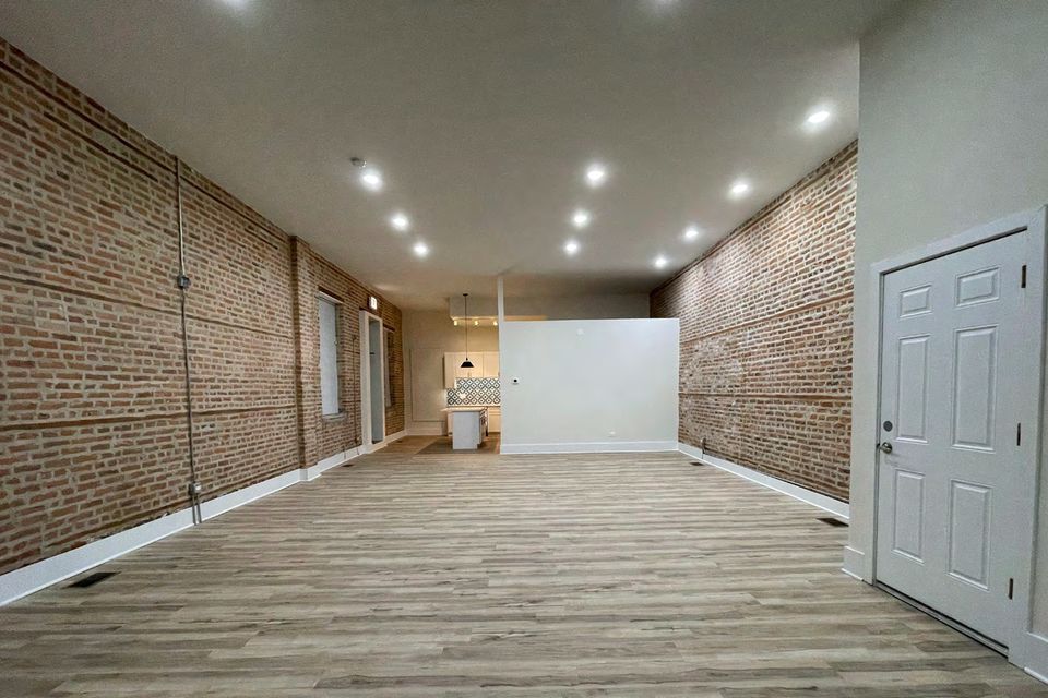 An empty room with an exposed brick wall, pale wood flooring and a row of ceiling lights.