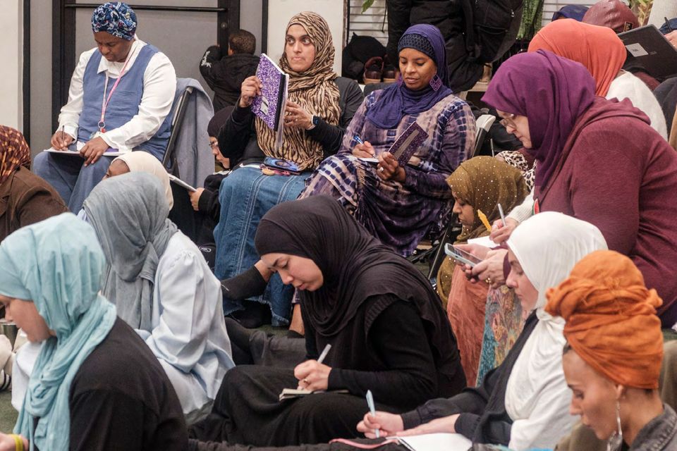 People seated in rows, writing in notebooks, in head coverings of many colors.