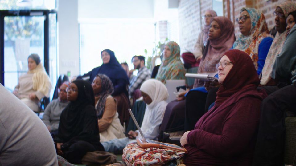 Women seated in rows indoors, listening, with daylight from a window at the left.