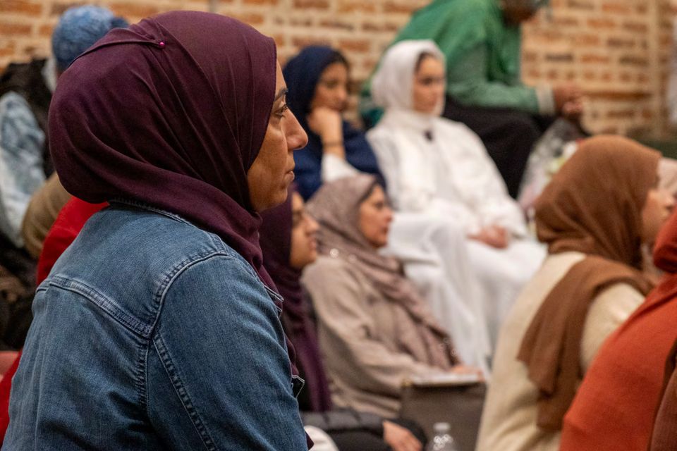 A woman in a maroon head covering seen in profile among seated people, against a brick wall.