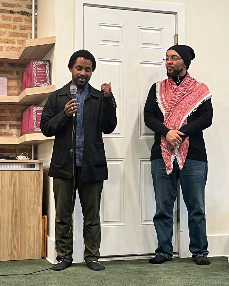 Two men standing side by side in front of a bookshelf, one holding a microphone.